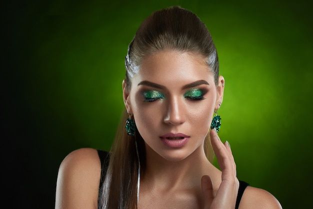 Premium Photo | Front view of sensual brunette girl with shiny green makeup touching perfect bronze skin of face. beautiful woman with closed eyes,wearing in black top, big rounded earring seductive posing .